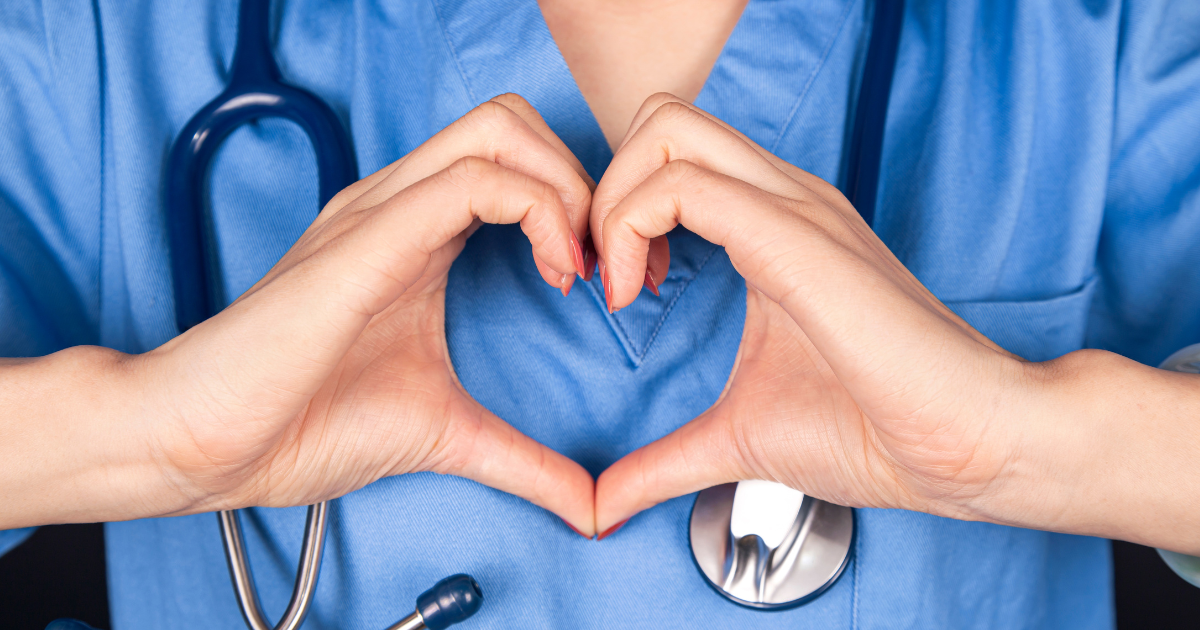 A close-up of a nurse forming a heart shape with their hands over blue scrubs and a stethoscope, symbolizing compassion and gratitude for Nurses Week.