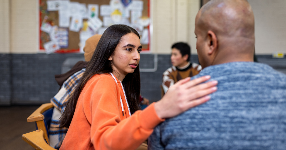 Woman in a support group gently placing her hand on another participant’s shoulder, symbolizing encouragement, empathy and community in addiction recovery.