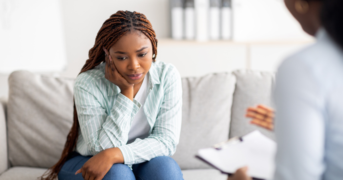Woman sitting on a couch with a thoughtful expression while speaking to a counselor, representing the challenges of stigma and the importance of supportive addiction treatment.