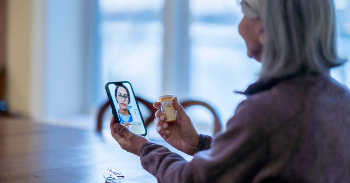 An older adult holding a smartphone during a telehealth visit while showing a prescription bottle, illustrating remote medication management in addiction recovery.