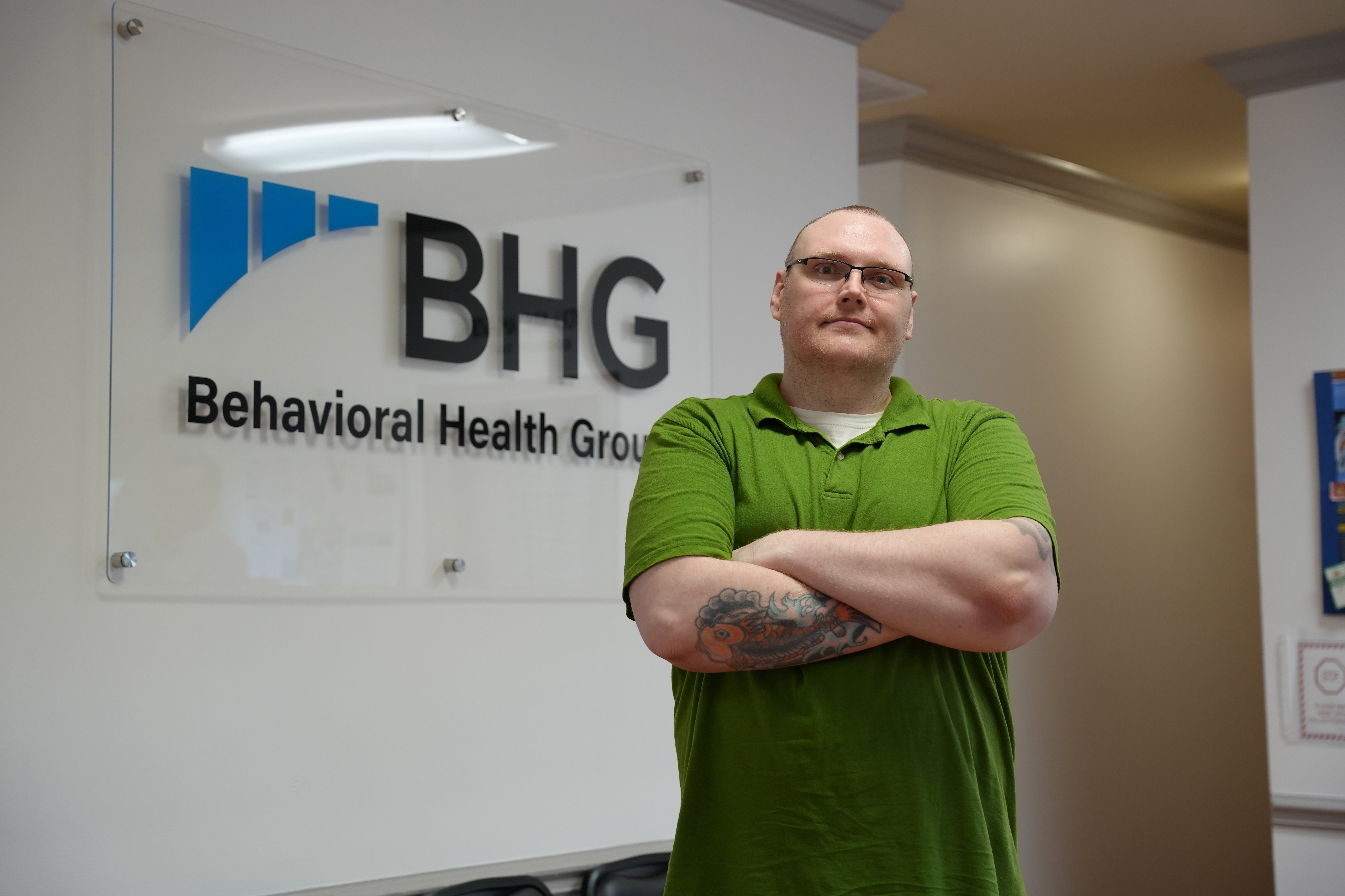 A man named Casey stands confidently inside a Behavioral Health Group center, arms crossed in front of a wall sign displaying the BHG logo. He appears calm and self-assured, representing strength and progress in his recovery journey from opioid use disorder.
