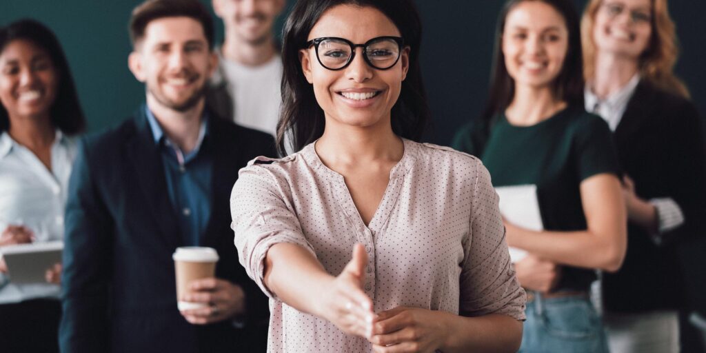 Woman in glasses extending hand for handshake with diverse team smiling behind her.