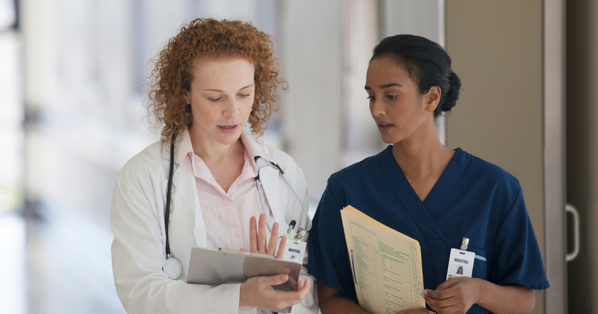 Two healthcare professionals walking and conversing in a clinical hallway - one wearing a white coat with a stethoscope, the other in scrubs holding a chart.