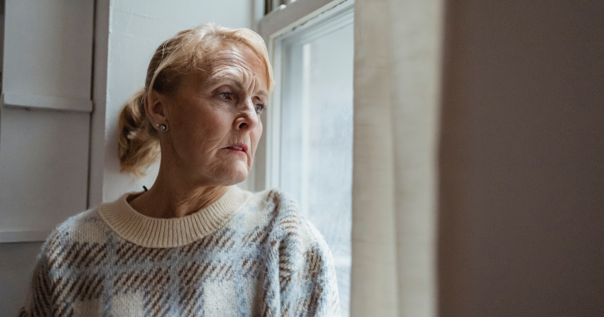 An older woman wearing a sweater looks thoughtfully out a window, reflecting concerns about potential Medicaid cuts and access to opioid treatment.
