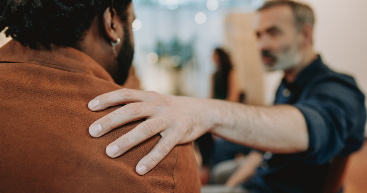 A support group session where one person places a reassuring hand on another's shoulder, symbolizing community, empathy and encouragement in recovery from opioid use disorder (OUD).
