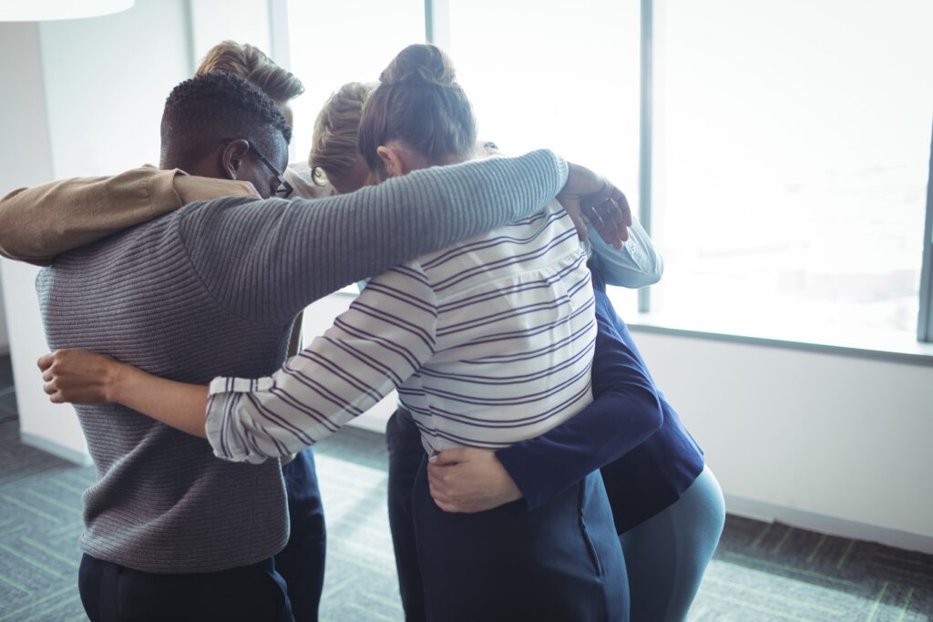 Four coworkers stand in a close huddle with their arms around each other in a bright office, heads bowed together, conveying teamwork, support, and collaboration.
