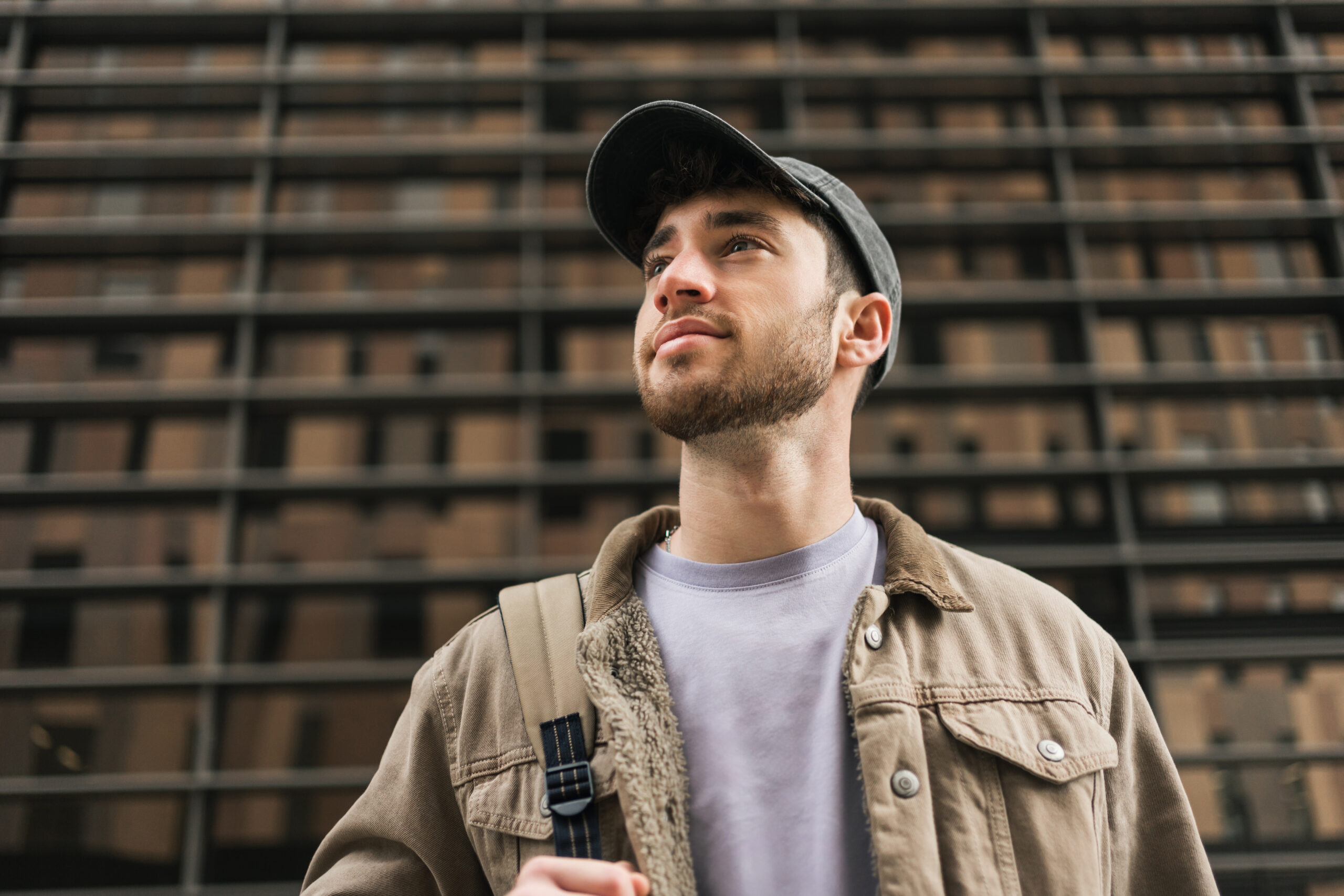 Young man in cap and tan jacket looking upward thoughtfully against urban building backdrop.
