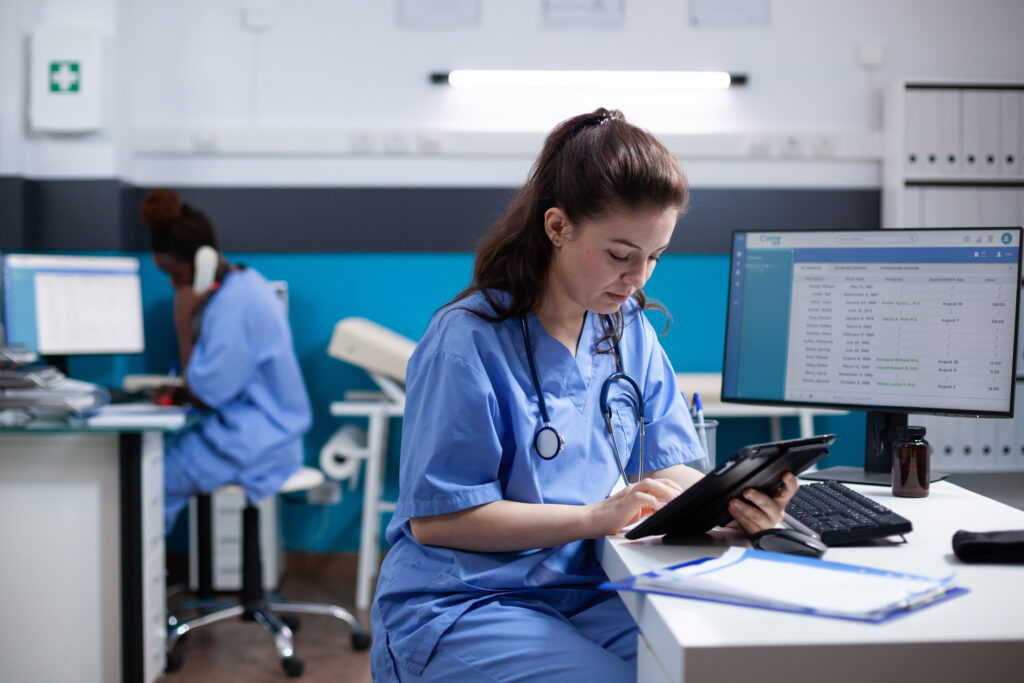 Healthcare professional in blue scrubs reviewing patient appointments on tablet in medical office.
