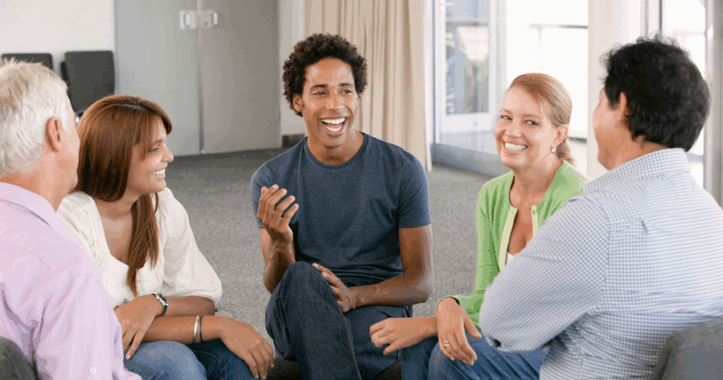 Diverse group of five people sitting in circle, smiling and engaged in conversation during support group meeting.