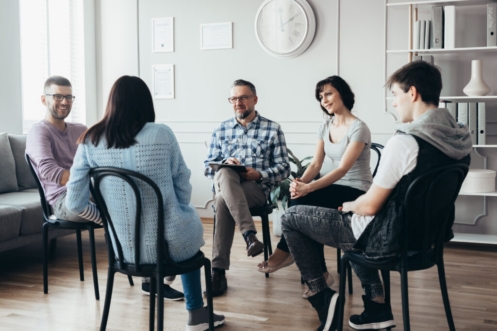 Five diverse people sitting in circle during supportive group counseling session in modern office.