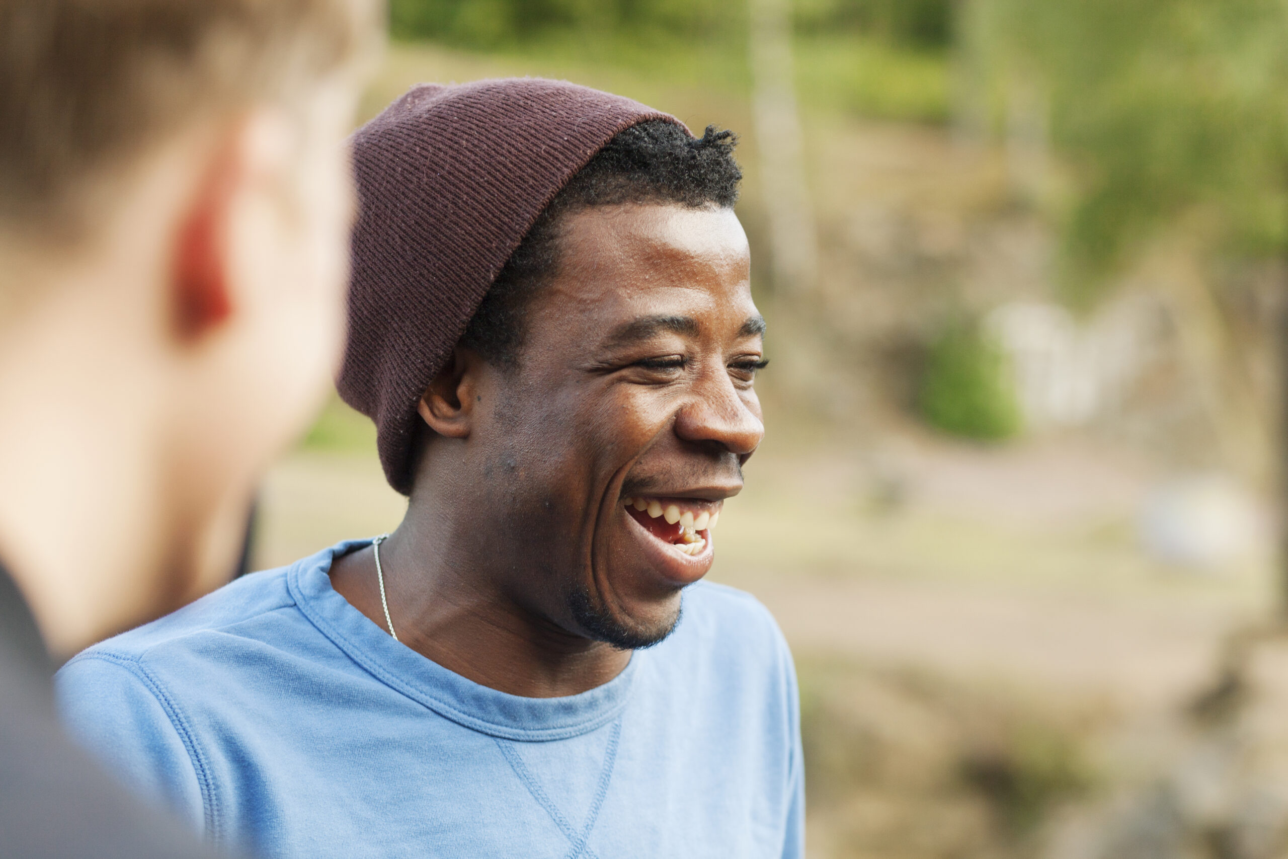 Smiling man in blue shirt and brown beanie laughing outdoors with friend blurred in background.