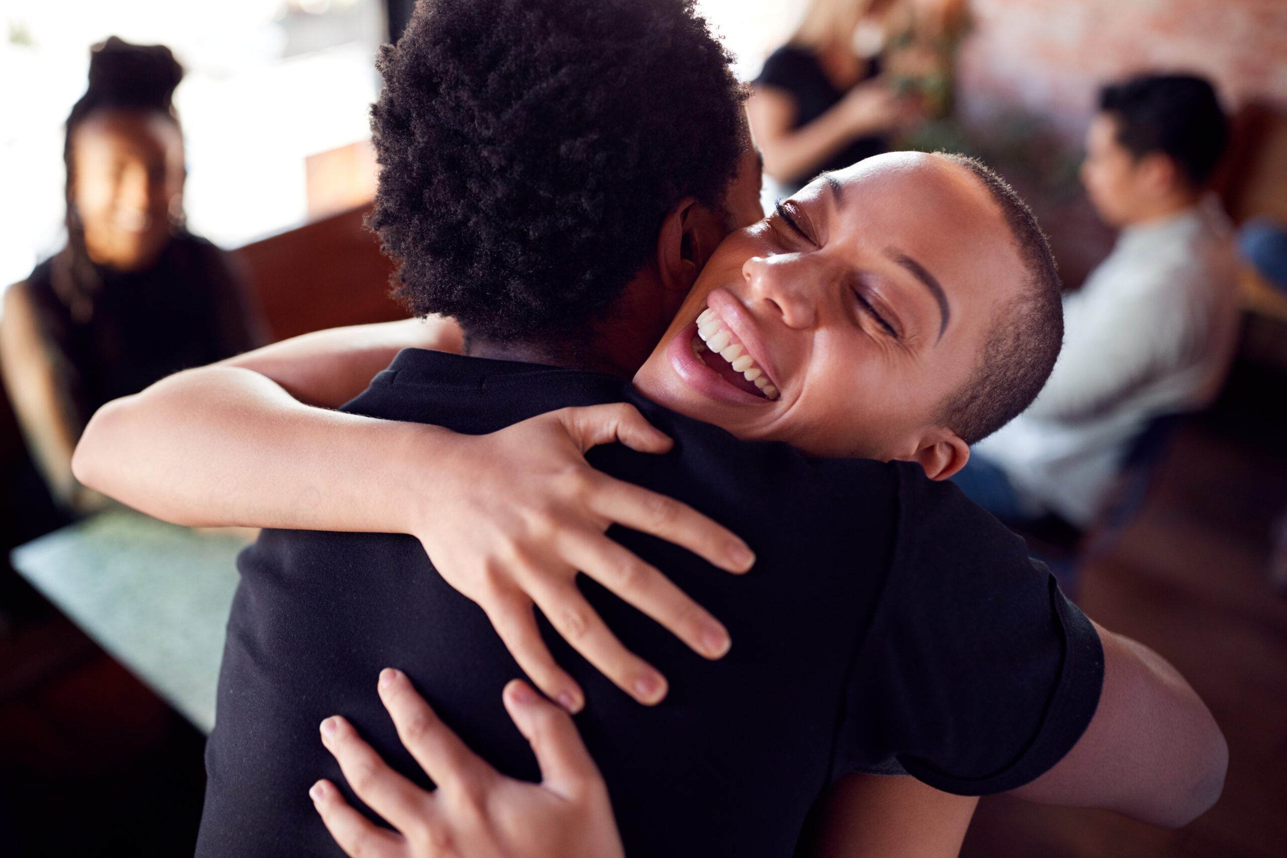 Two people embracing warmly with genuine smiles in a social setting, symbolizing connection and recovery support.