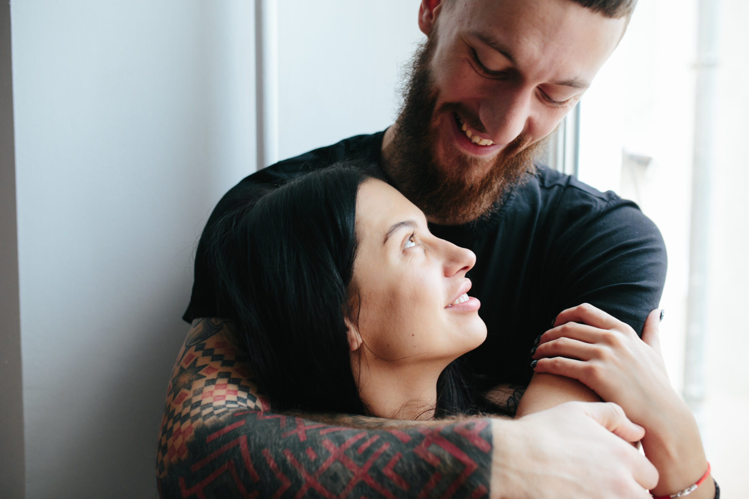Bearded man embracing woman from behind by window, both smiling joyfully together.