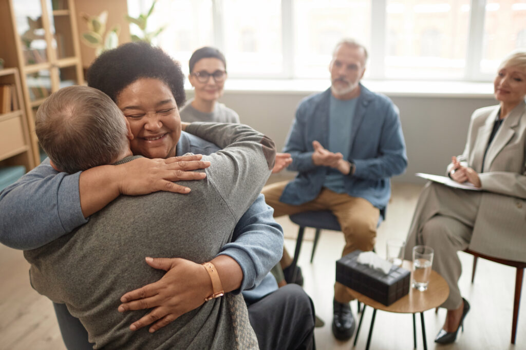 Senior woman hugging friend during group therapy session with diverse participants watching.
