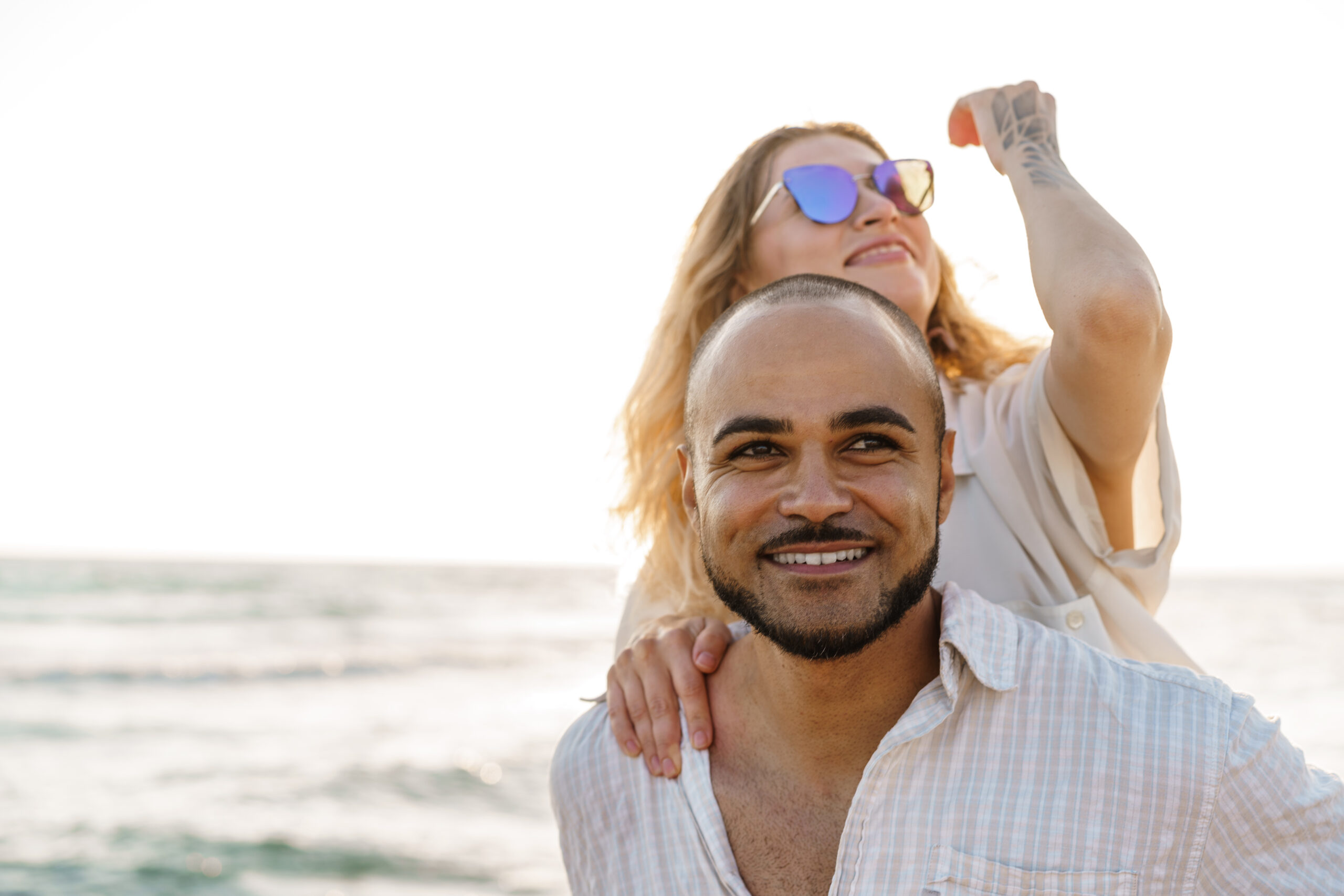 Happy couple embracing on beach, man smiling at camera, woman in blue sunglasses pointing ahead.