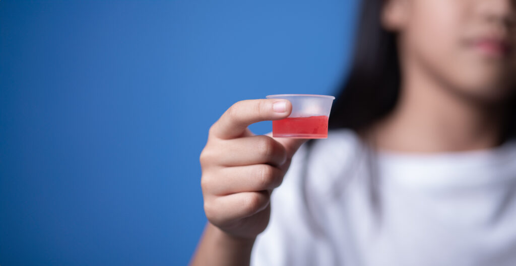 Child holding small cup of red liquid medicine against blue background, healthcare concept.