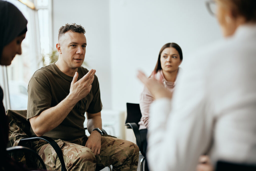 Veteran in military uniform speaking during a group therapy session with a counselor and other participants.