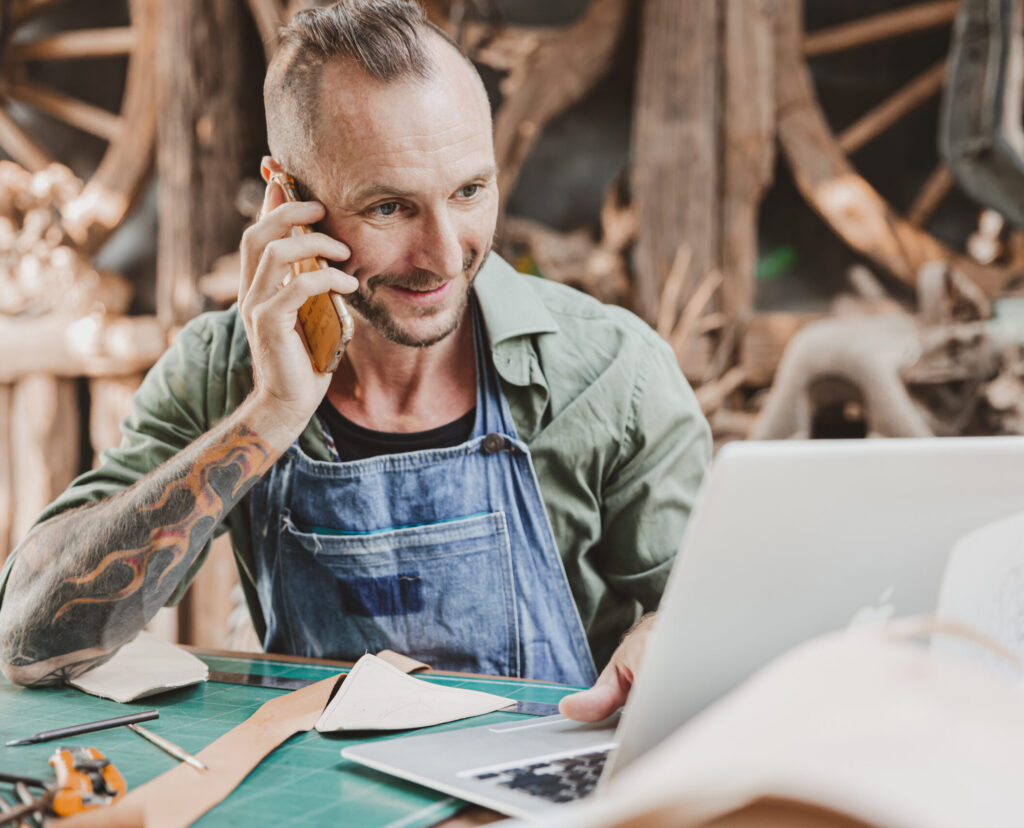 A man in a workshop smiling while talking on the phone and working at a laptop.